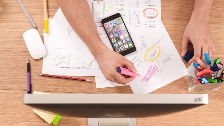 schwarzes iphone im homescreen auf beschriebenem papier auf tisch mit stift und monitor