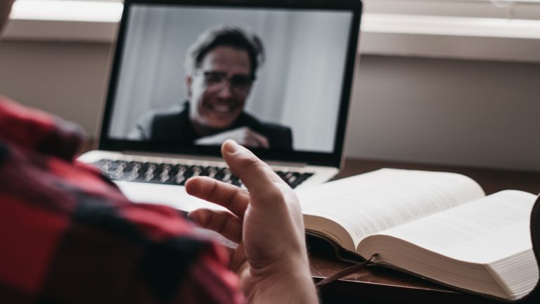Person mit rot-schwarz gestreiftem Hemd vor schwarzem Laptop und Buch